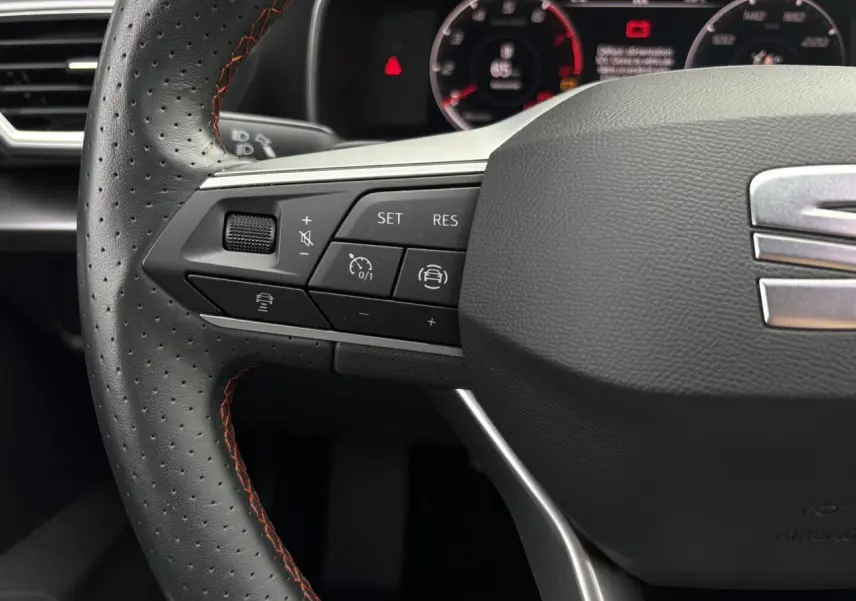 Gros plan sur le volant cuir noir perforé avec surpiqûres rouges du SEAT LEON 2024, boutons de régulateur de vitesse visibles.