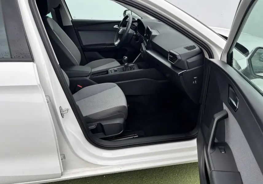 Intérieur côté conducteur d'une SEAT Leon blanche 2024, montrant sièges gris, volant cuir et tableau de bord moderne.