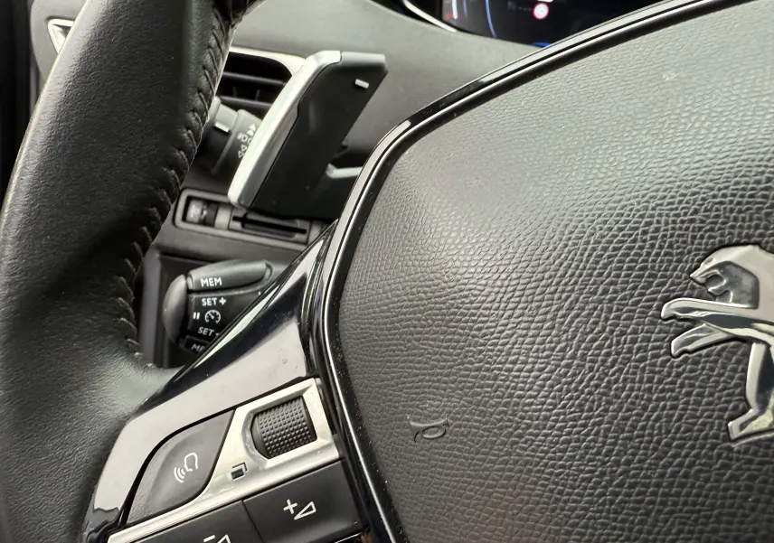 Gros plan sur le volant en cuir noir du Peugeot 5008 gris Artense, avec commandes intégrées et logo lion visible.