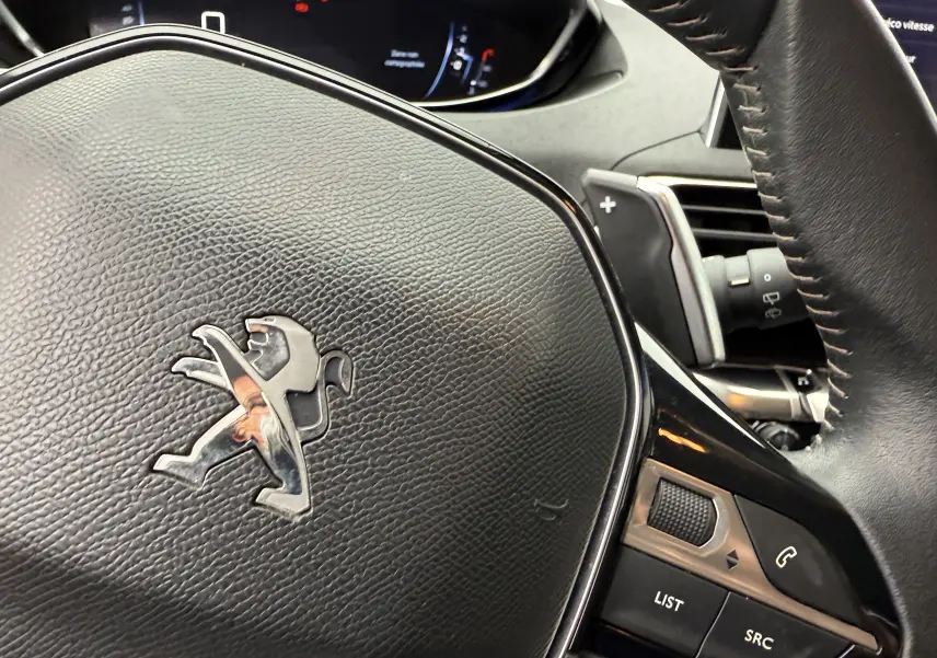 Gros plan sur le volant en cuir noir du Peugeot 5008, avec logo lion argenté et commandes intégrées visibles.