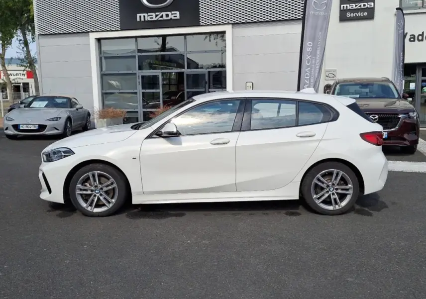 Profil côté gauche d'une BMW Série 1 blanche 120i M Sport 2021, avec jantes aluminium et lignes sportives visibles.