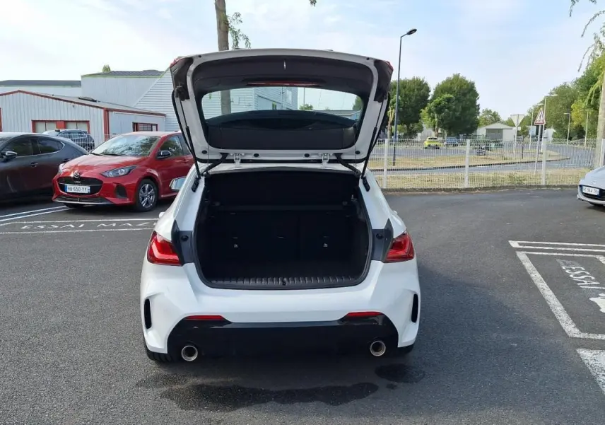 Vue arrière d'une BMW Série 1 blanche 120i M Sport 2021 avec coffre ouvert sur un parking extérieur.