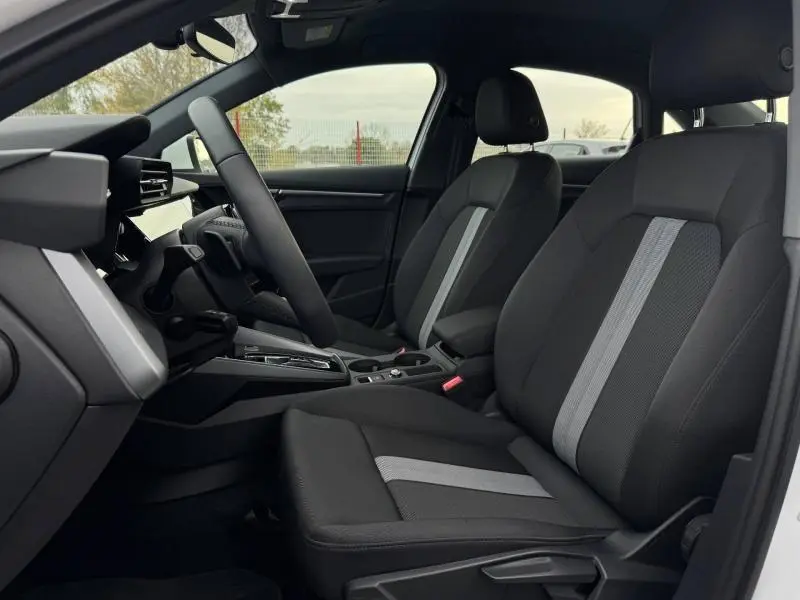 Vue intérieure côté gauche de l'Audi A3 Sportback 2024, sièges tissu noir avec bande grise centrale et volant cuir multifonction.