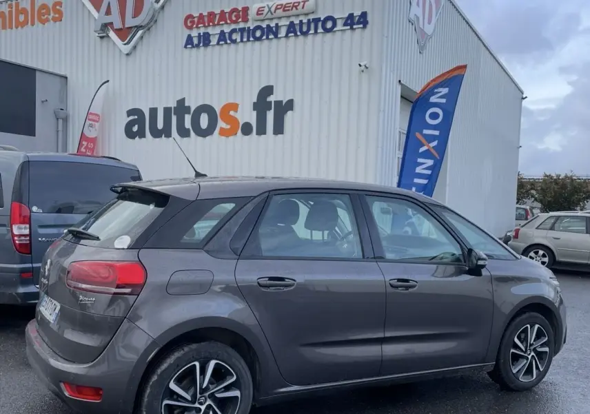 Vue côté gauche arrière d'une Citroën C4 Picasso marron avec jantes alu, stationnée devant un garage autos.fr par temps couvert.