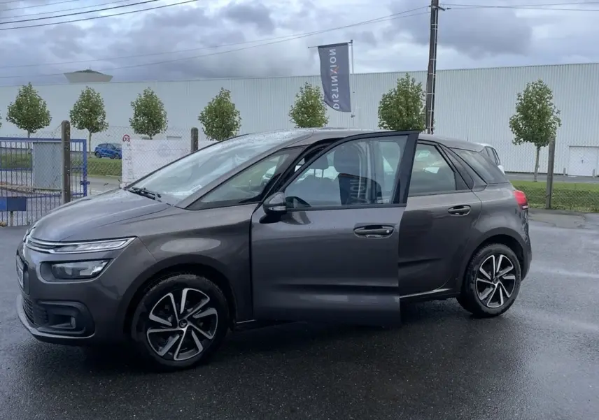 Vue de profil côté gauche d'une Citroën C4 Picasso marron avec porte avant ouverte et jantes alu sur sol humide.