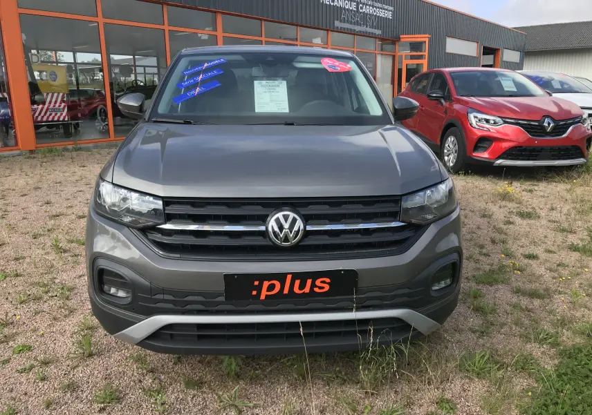 Vue de face d'un Volkswagen T-Cross gris limestone 2020 avec plaques :plus et autocollants promotionnels sur le pare-brise.