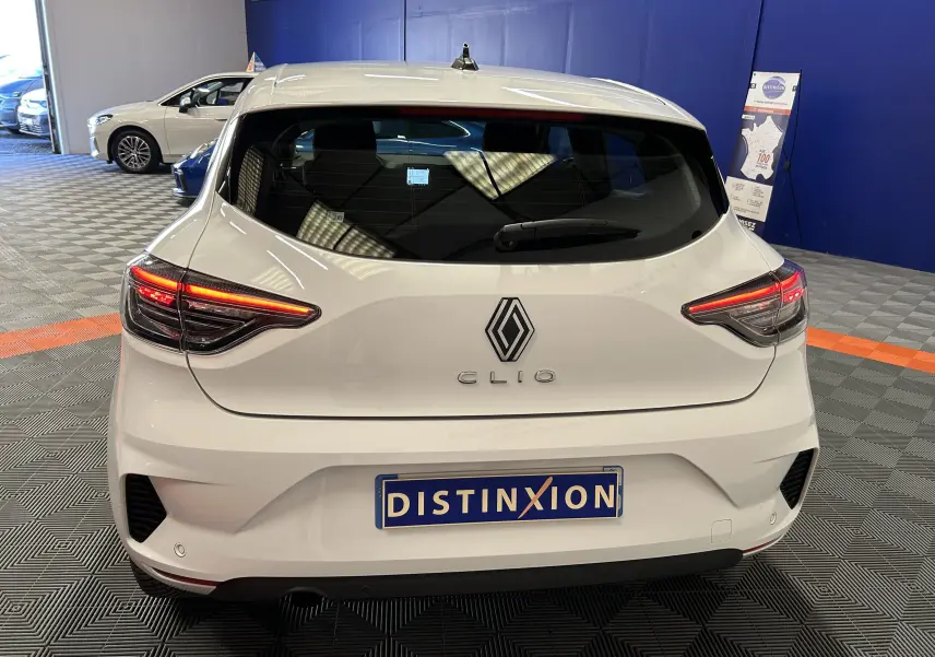 Vue arrière d'une Renault Clio V blanche avec feux arrière allumés dans un showroom au sol gris et murs bleus.
