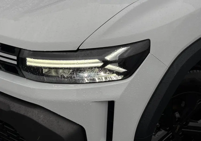 Gros plan sur le phare avant gauche du Dacia Duster blanc Glacier 2026 avec jante noire et gouttes de pluie.
