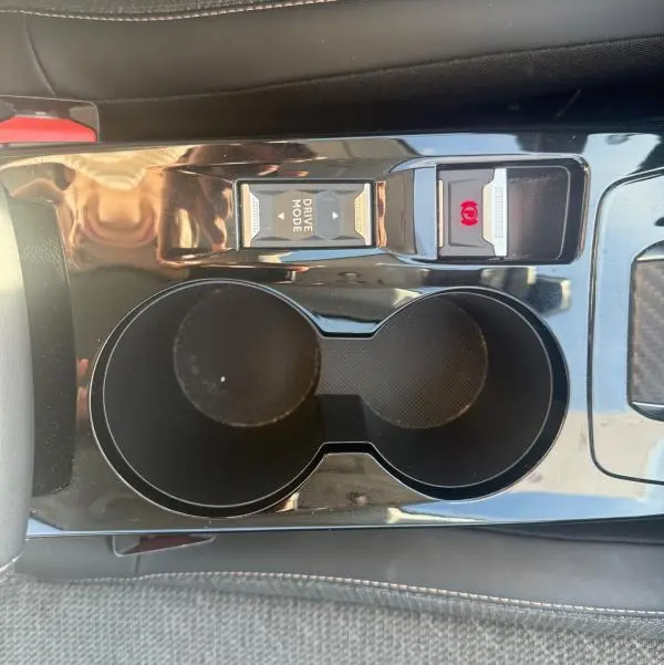 Gros plan sur la console centrale noire de la Peugeot 208 Hybrid 110 e-DCS6 Allure 2024, avec porte-gobelets et bouton Drive Mode.