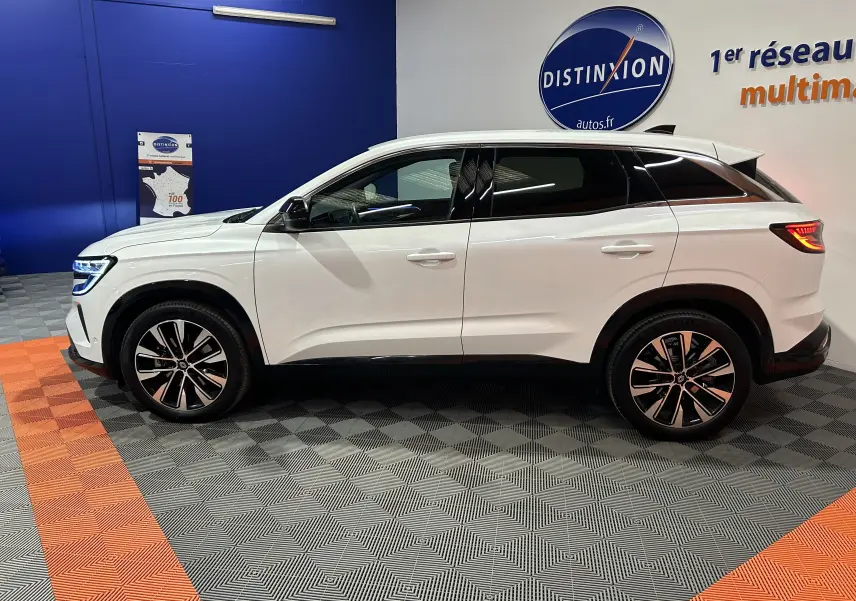 Profil droit du Renault Austral blanc nacré 2024 en hybride, avec jantes alliage et hayon électrique visible en arrière-plan.