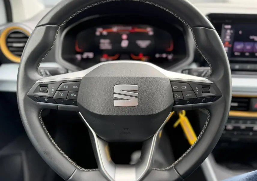 Vue rapprochée du volant noir du SEAT Arona 2025 avec commandes intégrées et tableau de bord digital en arrière-plan.
