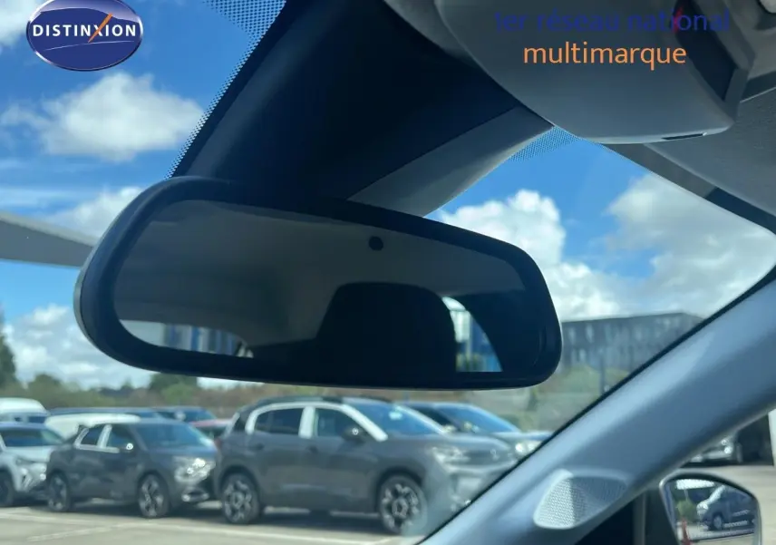 Rétroviseur intérieur reflétant le profil droit d'une Opel Corsa gris kristal métal stationnée en extérieur sous un ciel partiellement nuageux.