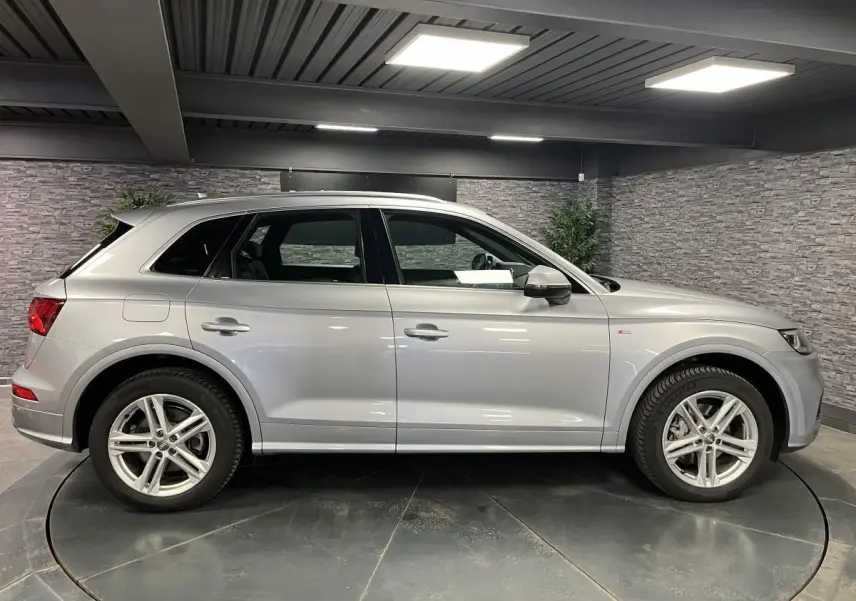 Audi Q5 gris en vue profil côté gauche dans un showroom, avec jantes alliage et badge S line visible sur l'aile avant.