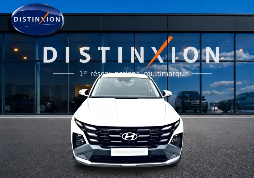 Vue frontale d’un Hyundai Tucson blanc 2026 avec calandre noire distinctive, devant une vitrine de concession multimarque.