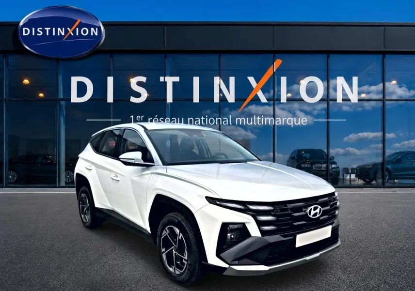 Vue 3/4 avant gauche d’un Hyundai Tucson blanc 2026 avec jantes alliage et calandre noire devant un bâtiment vitré Distinxion.