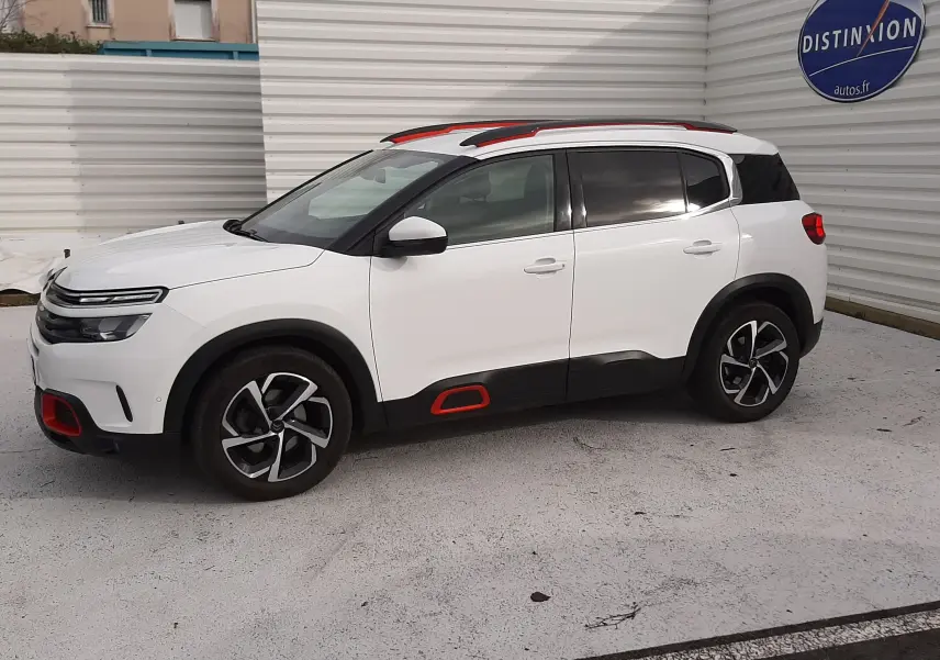 Vue côté gauche en 3/4 avant d'un Citroën C5 Aircross blanc avec détails rouges et toit noir, stationné devant un mur blanc.