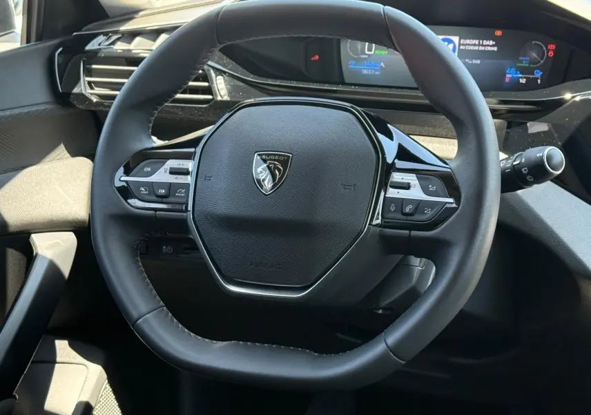 Vue rapprochée du volant multifonction noir avec logo Peugeot au centre, tableau de bord numérique en arrière-plan.