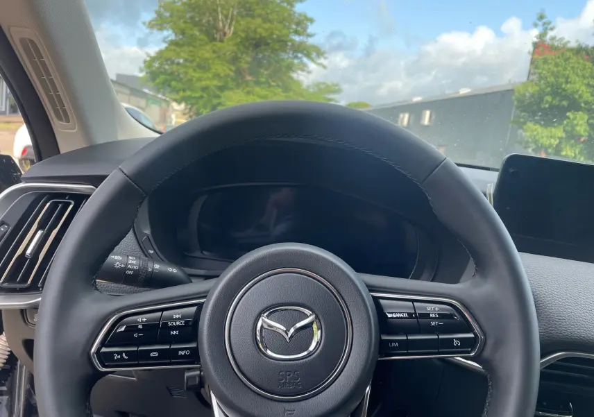 Vue rapprochée du volant en cuir noir du Mazda CX-60 2024, avec commandes intégrées et tableau de bord numérique éteint.