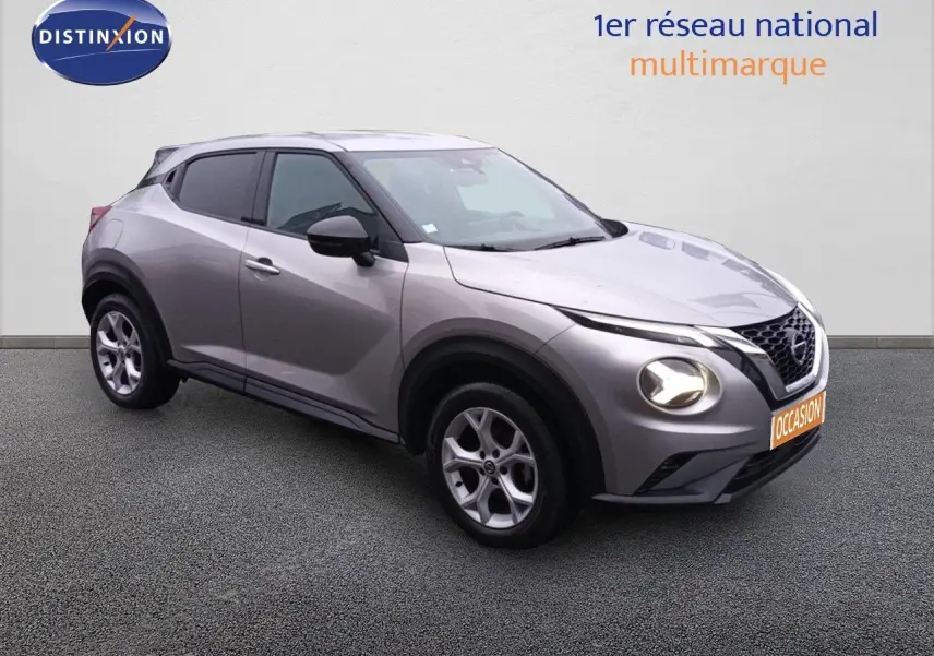 Nissan Juke gris platine métal en 3/4 avant droit, avec phares allumés et jantes alliage visibles.