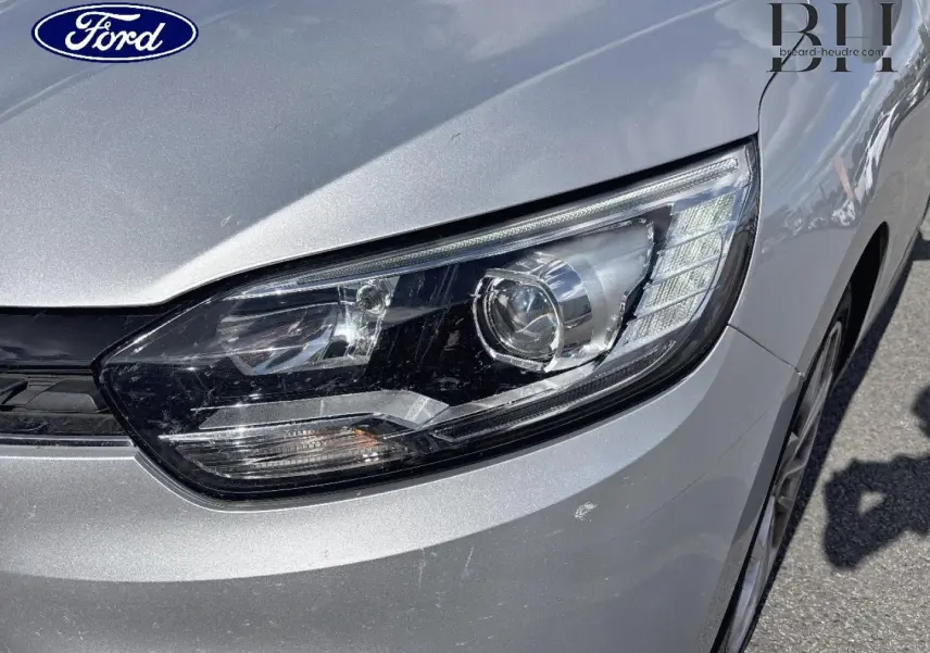 Gros plan sur le phare avant droit d'un Renault Grand Scenic gris platine, avec détails du capot et pare-chocs visibles.