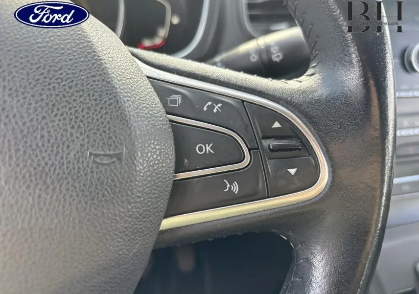 Gros plan sur le volant en cuir noir avec commandes intégrées, logo Ford visible en haut à gauche.