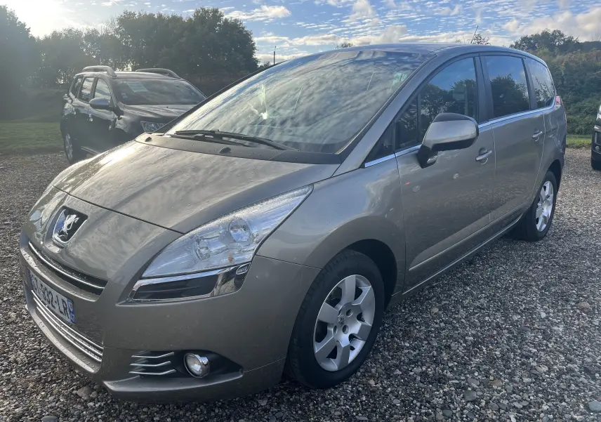 Peugeot 5008 2013 gris métallisé vue 3/4 avant droit sur un sol gravillonné avec ciel nuageux.
