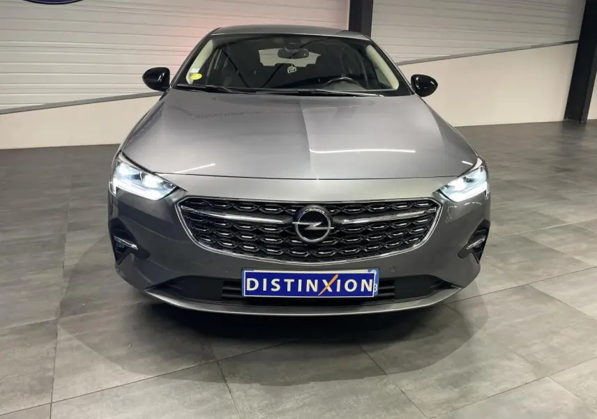 Opel Insignia Grand Sport gris clair vue de face avec calandre noire et phares LED allumés en intérieur showroom.