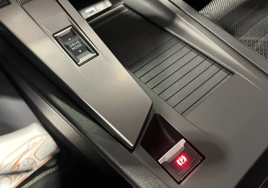 Gros plan sur la console centrale intérieure de la Peugeot 308 HYBRID 145 GT avec bouton Drive Mode et frein à main électronique rouge.