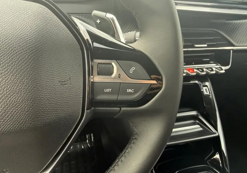Gros plan sur le volant noir de la Peugeot 208 Hybrid 145 e-DCS6 Allure 2025 avec commandes intégrées et tableau de bord brillant.