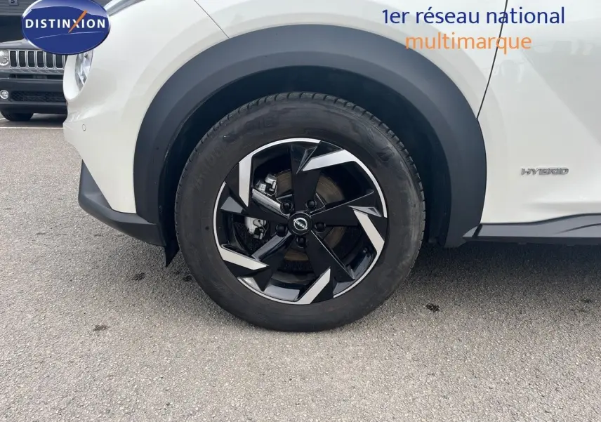 Gros plan sur la roue avant gauche noire et argentée du Nissan Juke blanc nacré hybride, avec badge Hybrid visible sur la carrosserie.