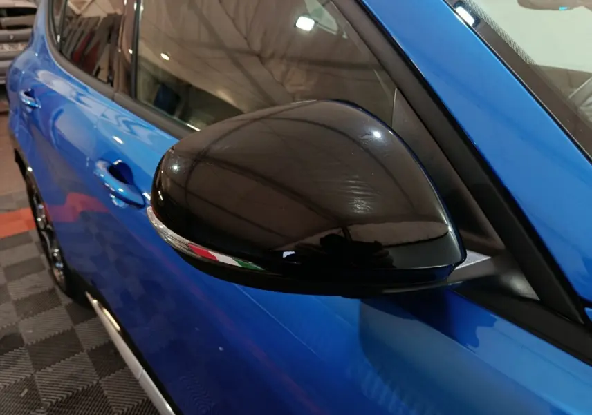 Gros plan sur le rétroviseur noir côté gauche de l'Alfa Romeo Tonale bleu, avec reflet du plafond de l'atelier.