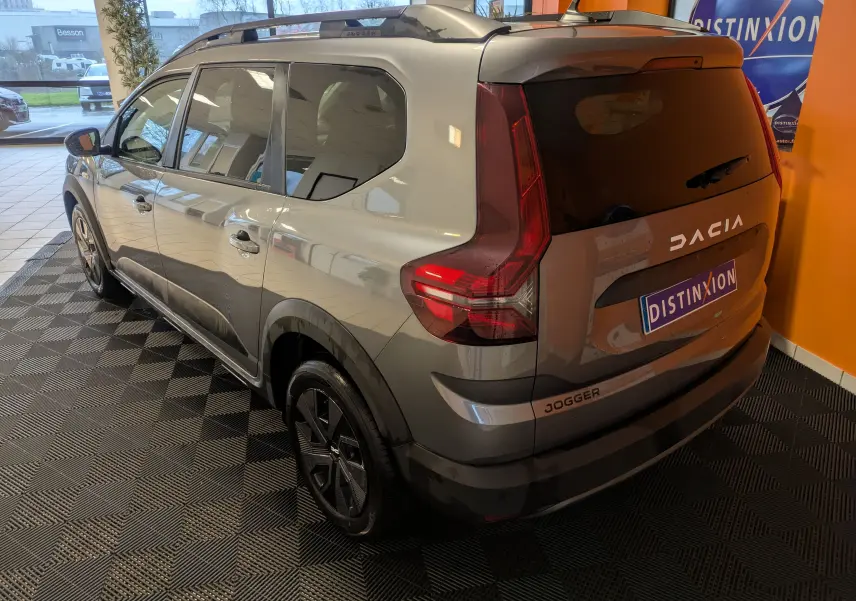 Vue 3/4 arrière droite du Dacia Jogger gris schiste métal avec feux arrière verticaux et jantes noires en intérieur showroom.