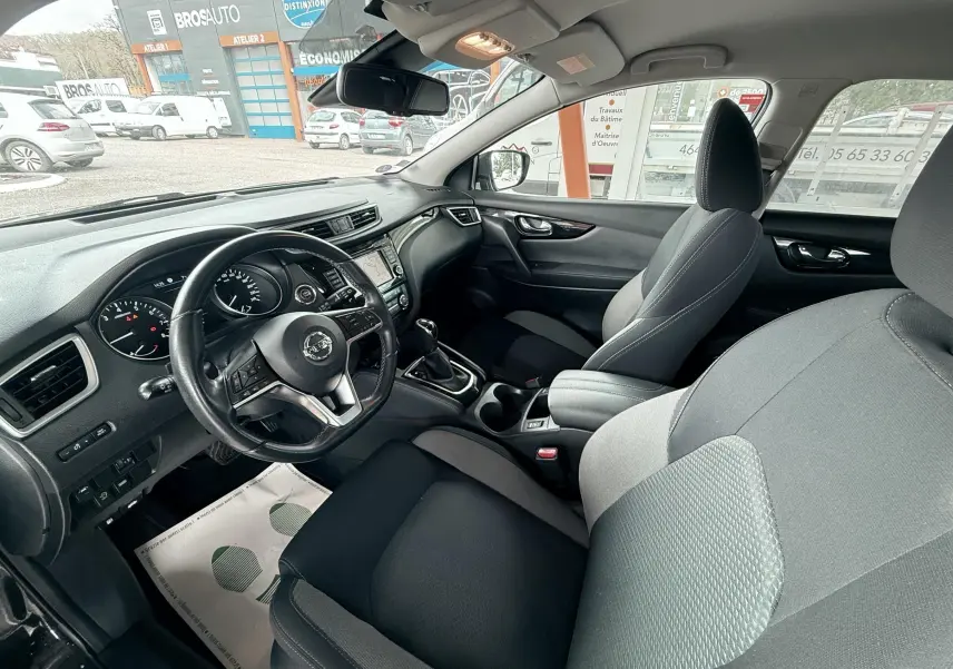 Vue intérieure côté conducteur du Nissan Qashqai 2020, montrant le volant, tableau de bord et sièges en tissu noir et gris.