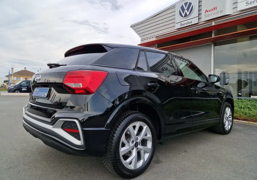 Audi Q2 noir mito metal vue 3/4 arrière droit, avec jantes alliage et vitres teintées, stationné devant un service Audi.
