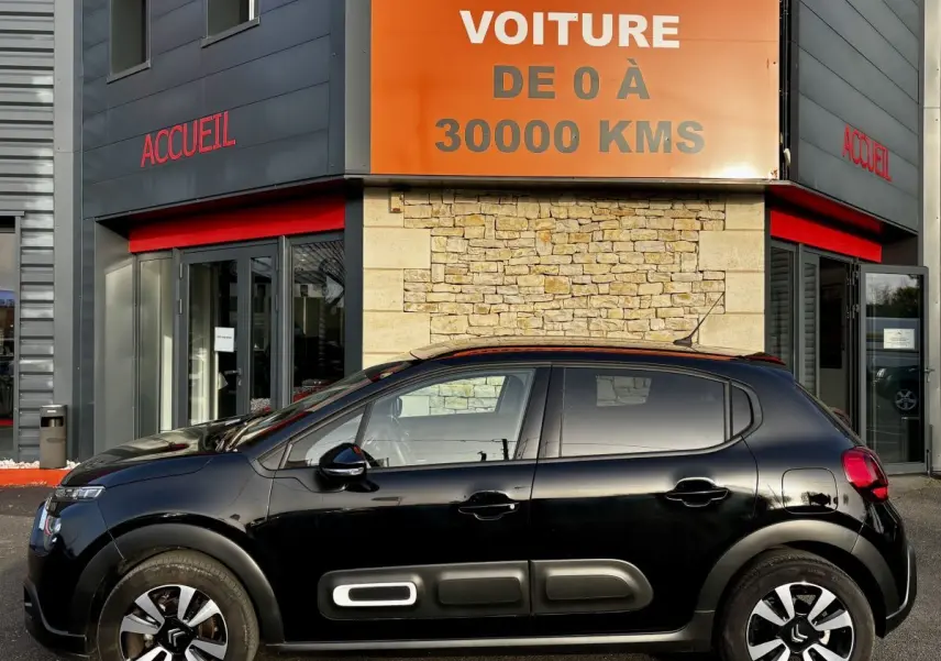 Profil latéral droit d'une Citroën C3 noire 2024 avec protections latérales blanches et jantes bicolores, devant un bâtiment commercial.