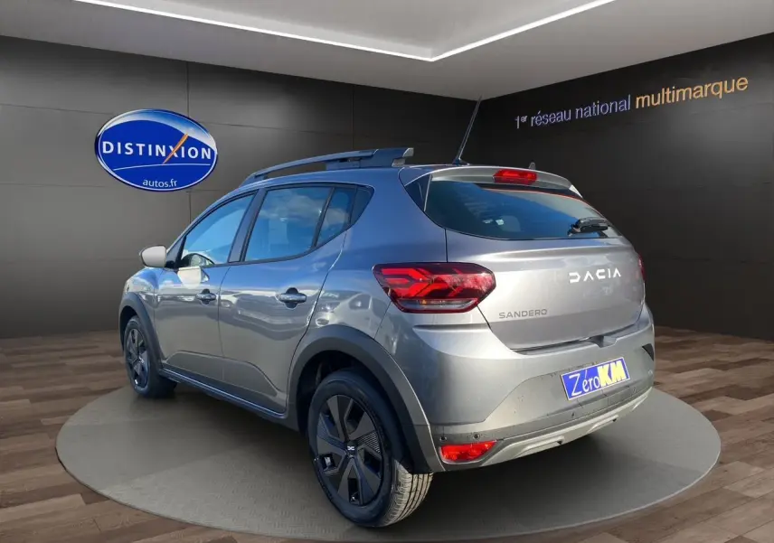 Vue 3/4 arrière droite d'une Dacia Sandero Stepway gris schiste avec barres de toit et jantes noires en showroom.