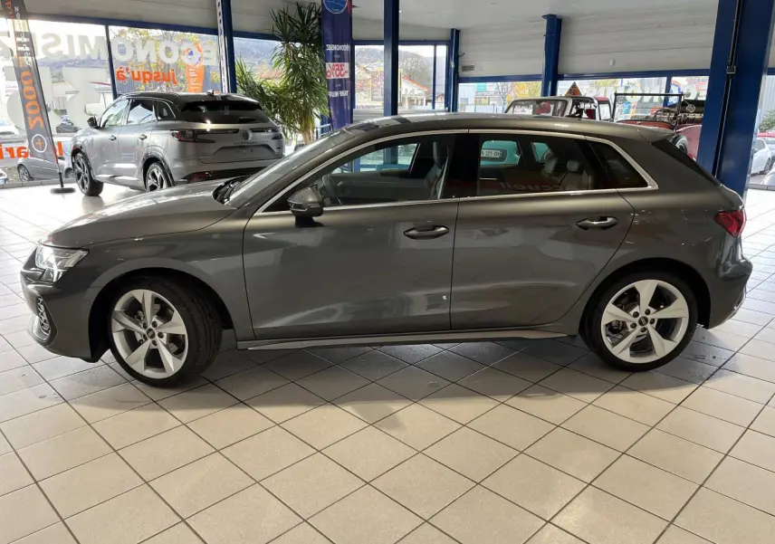 Audi A3 Sportback gris en profil droit dans un showroom, avec jantes alliage cinq branches et vitres teintées.