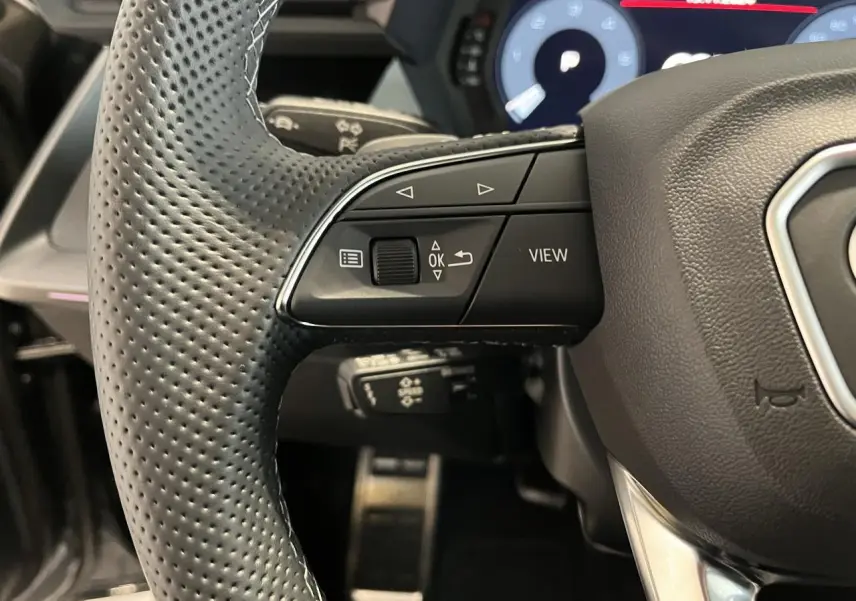 Gros plan sur le volant cuir perforé multifonctions de l'Audi A3 Sportback 2025 avec commandes intégrées et tableau de bord numérique.