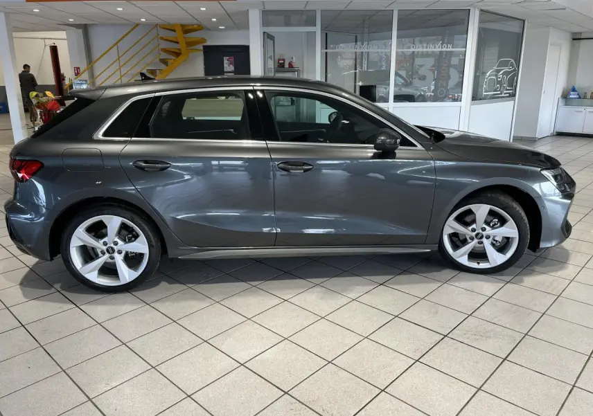 Vue de profil côté gauche d'une Audi A3 Sportback gris métallisé exposée en showroom intérieur.
