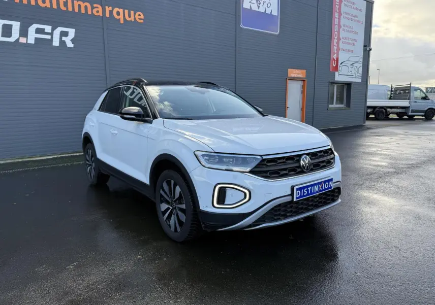 Volkswagen T-Roc blanc pur en 3/4 avant droit, avec jantes alliage et calandre noire devant un bâtiment gris.