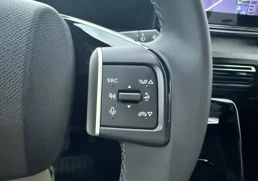 Gros plan sur les commandes gauche du volant noir de la Citroën C3 2025, avec tableau de bord et écran tactile en arrière-plan.