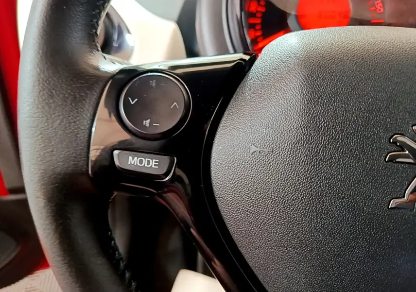 Gros plan sur le volant cuir multifonctions de la Peugeot 108 rouge, avec commandes audio intégrées et tableau de bord rouge en arrière-plan.