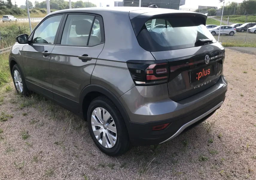 Vue 3/4 arrière droite d'un Volkswagen T-Cross gris Limestone avec feux arrière noirs et jantes argentées sur terrain gravel.