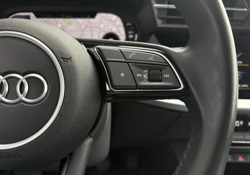 Gros plan sur le volant cuir noir multifonctions de l'Audi A3 Berline 35 TFSI 150 Design avec écran GPS en arrière-plan.