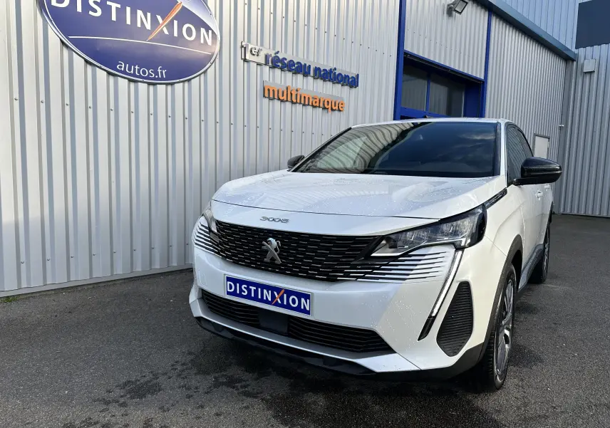 Peugeot 3008 blanc nacré en 3/4 avant droit, avec calandre noire et phares LED distinctifs, devant un bâtiment gris.