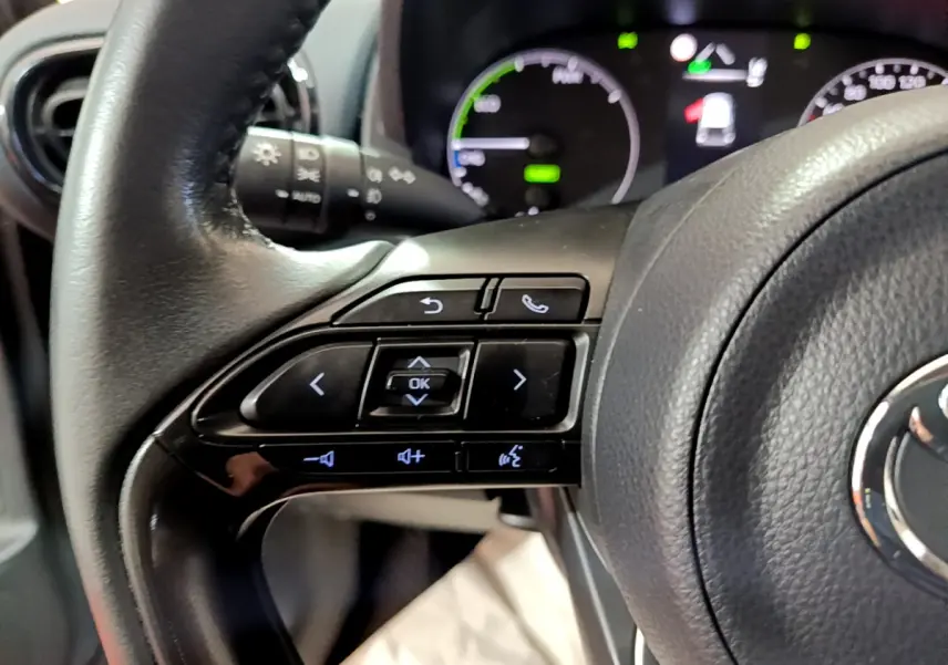 Gros plan sur les commandes du volant cuir de la Toyota Yaris hybride 2021, avec tableau de bord numérique en arrière-plan.