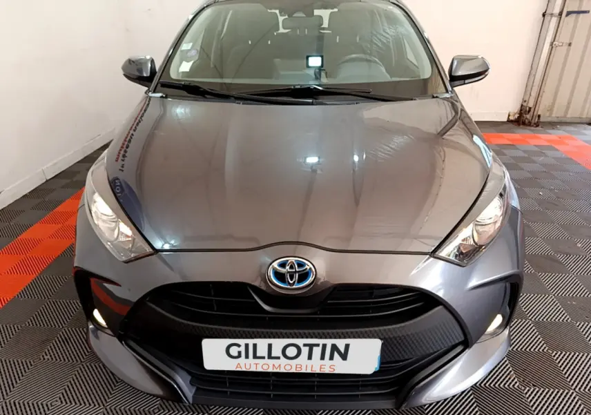 Vue frontale d'une Toyota Yaris hybride gris clair avec phares allumés dans un garage.