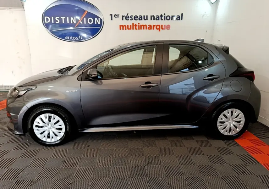 Profil côté gauche d'une Toyota Yaris hybride gris clair 2021, avec jantes à enjoliveurs blancs, en intérieur showroom.