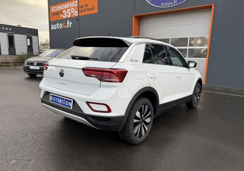 Vue 3/4 arrière droite d'un Volkswagen T-Roc blanc pur avec jantes alliage Zürich, stationné devant un garage autos.fr.