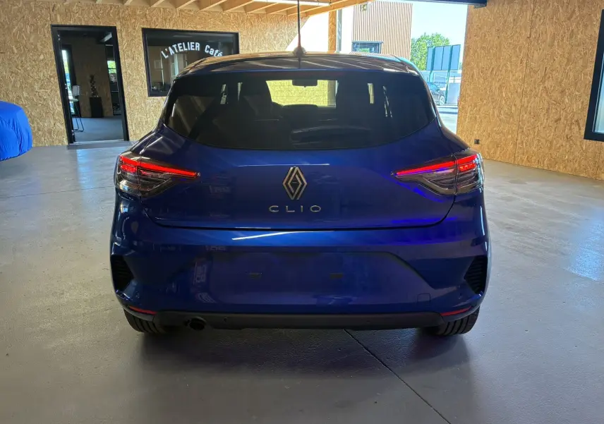 Vue arrière d'une Renault Clio 2024 bleu Iron, avec feux arrière allumés dans un intérieur d'atelier.